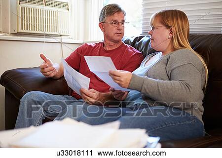 Stock Image - Worried Couple Sitting On Sofa Looking At Bills. Fotosearch