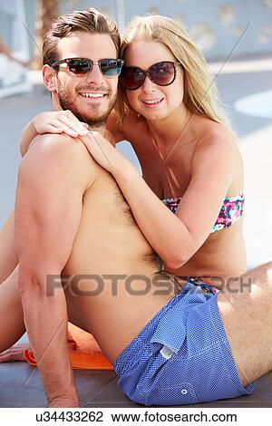 Stock Image - Young Couple On Holiday Relaxing By Swimming Pool. Fotosearch