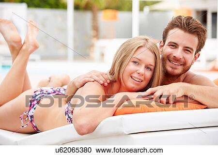 Stock Image - Young Couple On Holiday Relaxing By Swimming Pool. Fotosearch