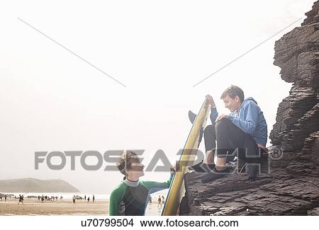Boy sitting on rocks with surfboard with his friend View Large Photo Image Picture - Boy sitting on rocks with surfboard with his friend. Fotosearch