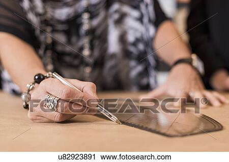 Stock Image - Close up of mature seamstress drawing with curved ruler on dressmakers pattern in workshop. Fotosearch