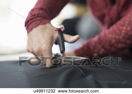 Stock Image - Close up of seamstress hands using scissors to cut textile at work table. Fotosearch