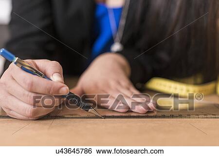 Stock Photograph - Close up of young seamstress drawing with ruler on dressmakers pattern in workshop. Fotosearch