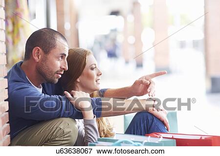 Stock Photo - Couple sitting by shopping bags, man pointing. Fotosearch