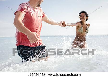 Stock Image - Couple wading and holding hands in sea, Tuscany, Italy. Fotosearch