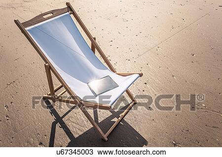 Stock Image - Deck chair on beach with electronic book on seat. Fotosearch