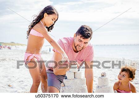 Stock Image - Female toddler and sister building sandcastle with father at beach, Tuscany, Italy. Fotosearch