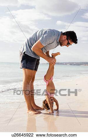 Stock Image - Female toddler doing handstand helped by father at beach, Tuscany, Italy. Fotosearch