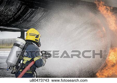 Stock Image - Fireman spraying water on simulated aircraft fire at training facility. Fotosearch
