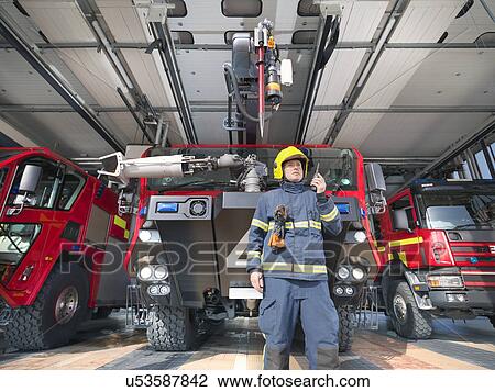 Stock Image - Fireman using walkie talkie in front of fire engines in airport fire station. Fotosearch