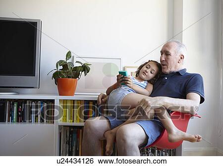 Girl sitting on grandfathers lap in living room playing hand held computer game View Large Photo Image Stock Photograph - Girl sitting on grandfathers lap in living room playing hand held computer game. Fotosearch