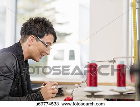 Stock Image - Mature seamstress picking sewing thread with tweezers in workshop. Fotosearch