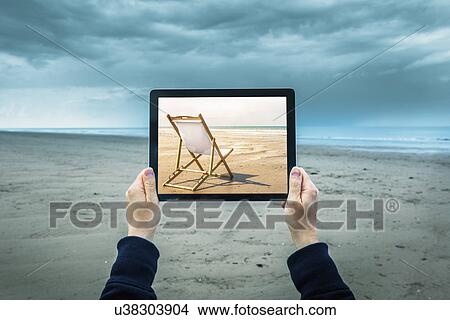 Picture - Mature woman on cloudy beach, holding digital tablet showing sunny beach scene, focus on hands and laptop. Fotosearch