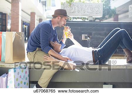 Stock Photography - Mid adult couple relaxing on seat, drinking drink, shopping bags beside them. Fotosearch