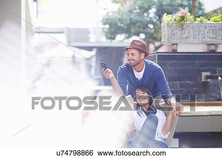 Stock Photograph - Mid adult couple sitting together outdoors, looking at smartphones. Fotosearch