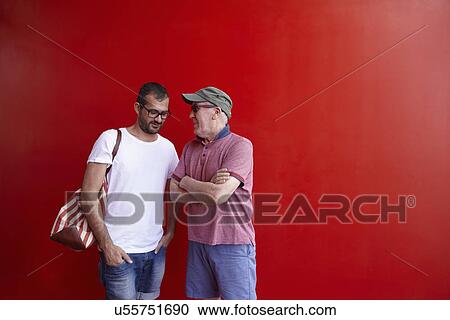 Stock Image - Mid adult man and senior man talking, standing against red background. Fotosearch