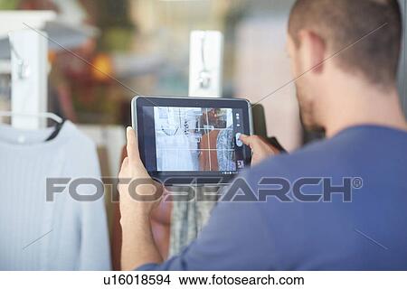 Picture - Mid adult man holding digital tablet, taking photograph of shop window. Fotosearch