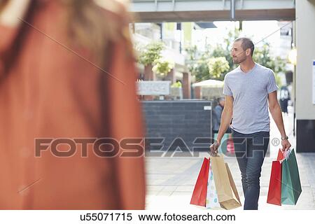 Mid adult man holding shopping bags, walking behind woman View Large Photo Image Stock Photography - Mid adult man holding shopping bags, walking behind woman. Fotosearch