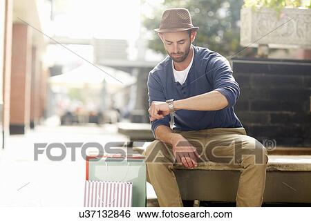 Stock Photograph - Mid adult man sitting on seat, looking at watch, shopping bags beside him. Fotosearch