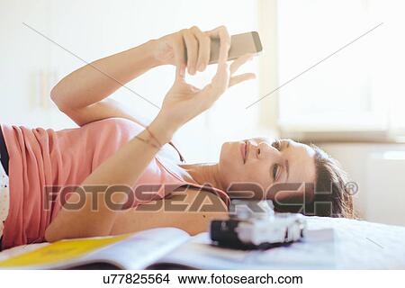 Picture - Mid adult woman lying on bed reading messages on smartphone. Fotosearch