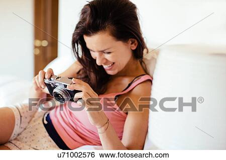 Stock Photograph - Mid adult woman lying on bed reviewing photographs on digital SLR camera. Fotosearch