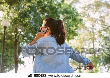 Stock Image - Mid adult woman pushing bicycle and talking on smartphone, Tuscany, Italy. Fotosearch