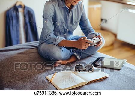 Stock Photograph - Mid adult woman sitting on bed reviewing photographs on digital SLR camera. Fotosearch