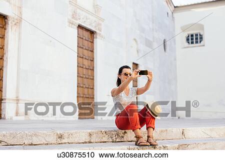 Stock Image - Mid adult woman sitting taking selfie in front of church with smartphone, Tuscany, Italy. Fotosearch