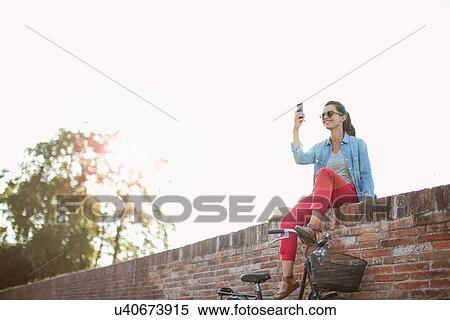 Stock Photography - Mid adult woman taking smartphone selfie on brick wall, Tuscany, Italy. Fotosearch