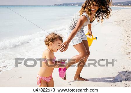 Stock Image - Mother and toddler daughter with toy watering cans on beach, Tuscany, Italy. Fotosearch