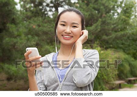 Stock Photography - Portrait of young female runner selecting music from smartphone in park. Fotosearch