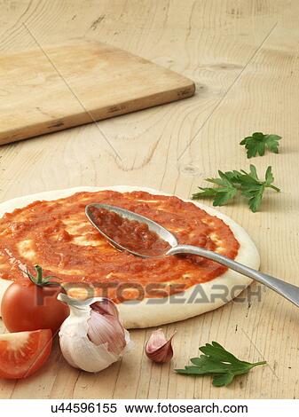 Stock Photography - Prepared pizza base covered in tomato sauce with garlic clove and chopped tomato. Fotosearch