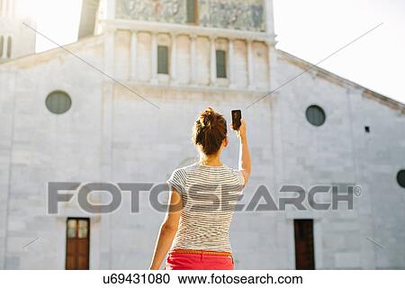 Stock Image - Rear view of mid adult woman photographing church with smartphone, Tuscany, Italy. Fotosearch