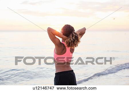 Stock Photo - Rear view of young woman stretching on beach at sunset, Tuscany, Italy. Fotosearch