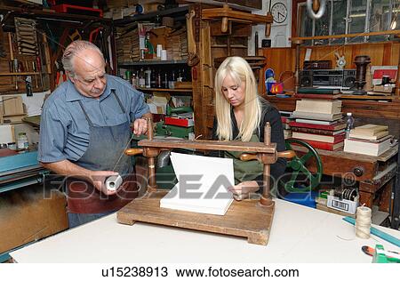 Stock Image - Senior man and young woman preparing to bind pages with thread in traditional bookbinding workshop. Fotosearch