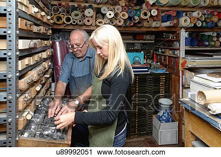 Stock Image - Senior man and young woman selecting book letterpress in traditional bookbinding workshop. Fotosearch
