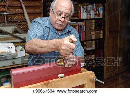 Picture - Senior man applying gold leaf to book spine in traditional bookbinding workshop. Fotosearch