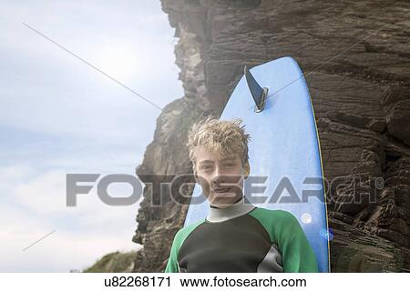 Stock Image - Teenage boy leaning against surfboard. Fotosearch
