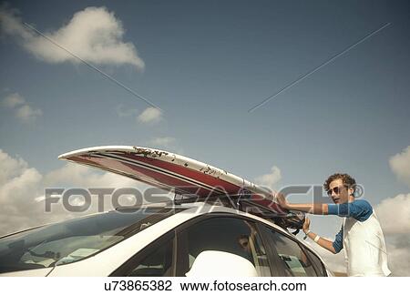 Stock Image - Teenage boy putting surfboard on top of car. Fotosearch