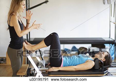 Stock Image - Tutor instructing female student lying on reformer in pilates gym. Fotosearch