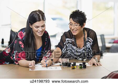 Stock Image - Two seamstresses painting fashion design on work table. Fotosearch