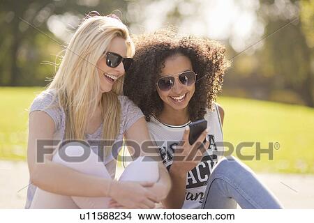 Two young female looking down at text message on smartphone in park View Large Photo Image Picture - Two young female looking down at text message on smartphone in park. Fotosearch