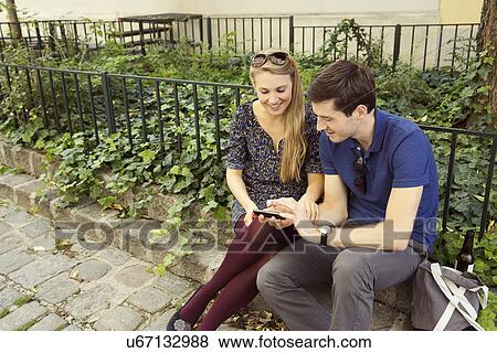 Young couple sitting on wall looking down at smartphone View Large Photo Image Stock Photo - Young couple sitting on wall looking down at smartphone. Fotosearch