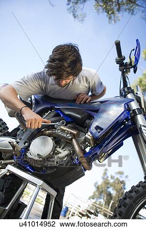 Stock Image - Young male motocross racer adjusting motorcycle engine in forest. Fotosearch