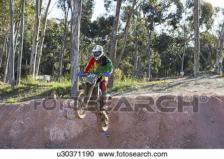 Stock Image - Young male motocross racer jumping down mud hill. Fotosearch