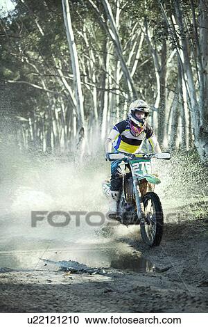 Stock Image - Young male motocross racer riding through puddle in forest. Fotosearch