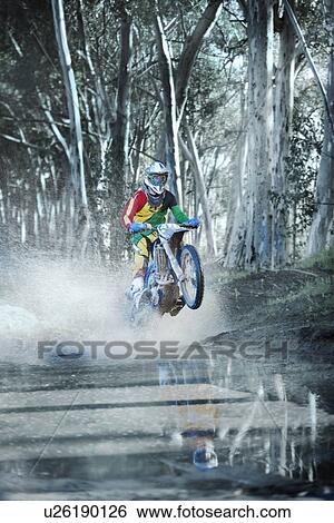 Stock Photograph - Young male motocross racer riding through dirt track puddle in forest. Fotosearch
