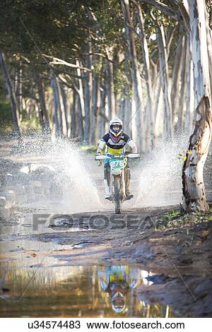Stock Image - Young male motocross racer splashing through puddle in forest. Fotosearch