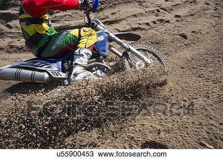 Stock Image - Young male motocross rider racing through mud track. Fotosearch
