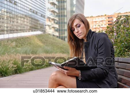 Picture - Young professional woman sitting on bench, looking in notebook. Fotosearch